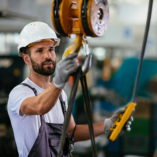 A crane operator wearing a hard hat holding a crane hook and a pendant controller in a manufacturing factory